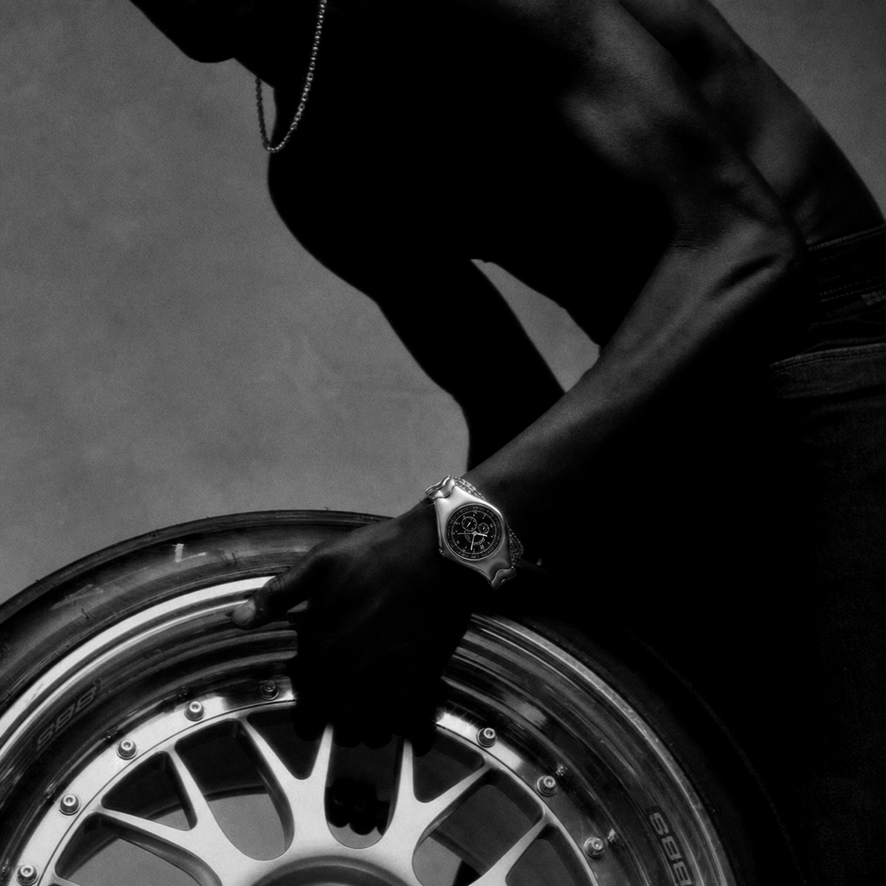 Photographie en noir et blanc d'une personne portant une montre-bracelet, la main posée sur une roue de voiture chromée.